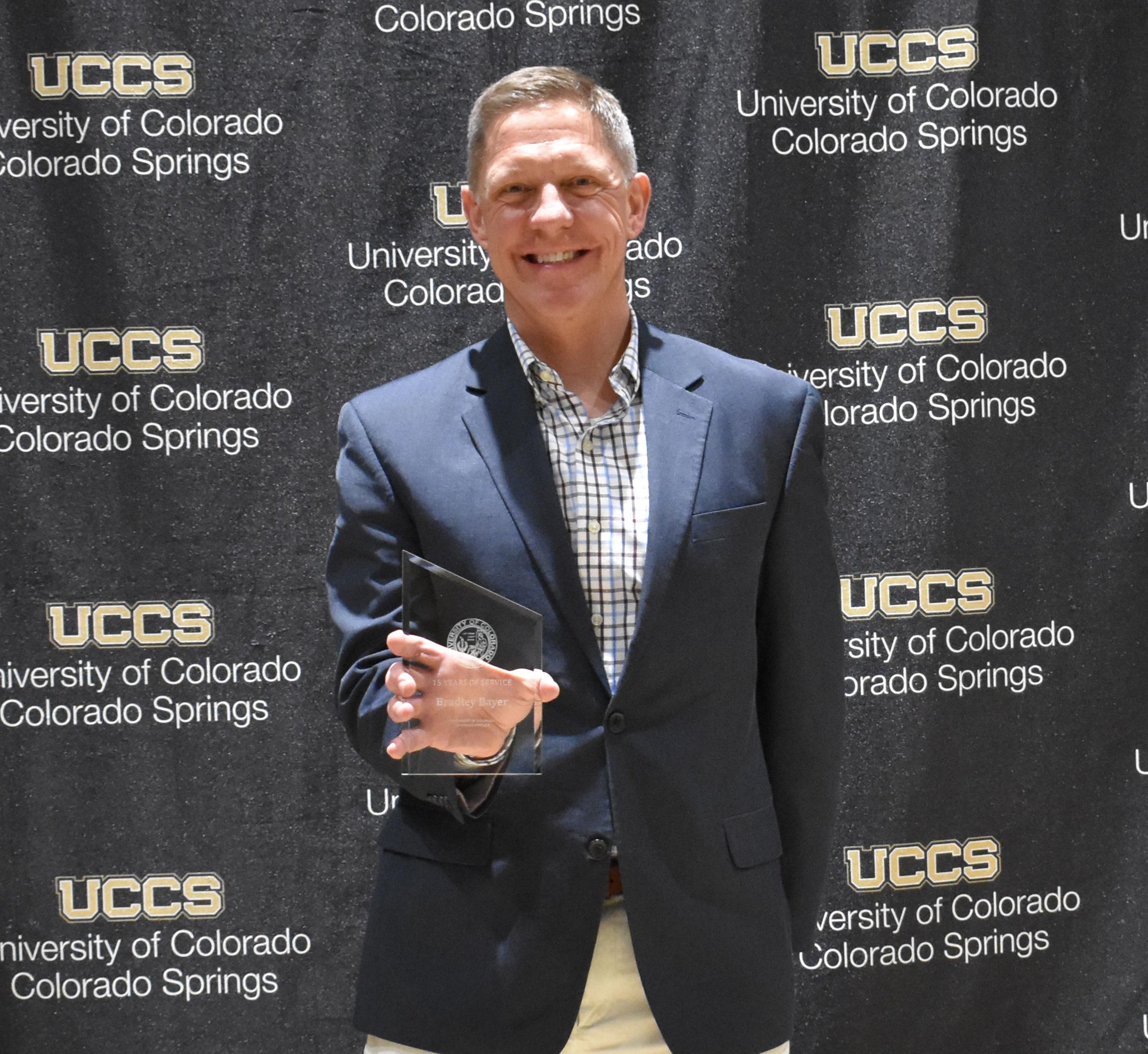 UCCS staff member holding his longevity award in front of a step and repeat that says UCCS