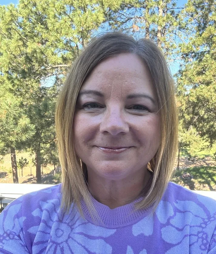 woman with should length hair in a purple floral sweater in front of some trees